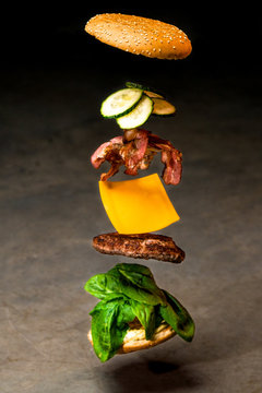 Flying cheeseburger ingredients on dark background of concrete. Fastfood concept. Ingredients of burger - bun, grilled cutlet, cheese, bacon, cucumber and spinach