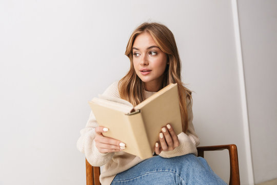 Image of thinking young woman reading book while sitting on armchair