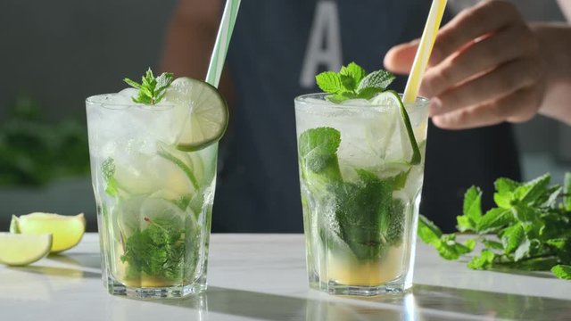 Barman puts on the bar table two mojito cocktails. Refreshing Mojito cocktail drinks in the glasses with fresh sliced limes and mint leaves. Party time. Summer mojito