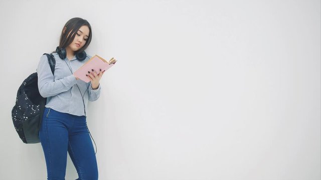 An asian schoolgirl with a backpack slung oveer shoulder, standing, reading a book, then waving hello someone.