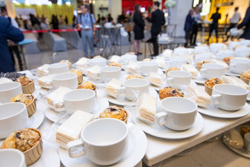 Group of empty coffee cups with snack cake on plate. Many rows of white cup for service hot tea or coffee in buffet and seminar event over Blurred people crowd backgrounds, shallow dof or more blur