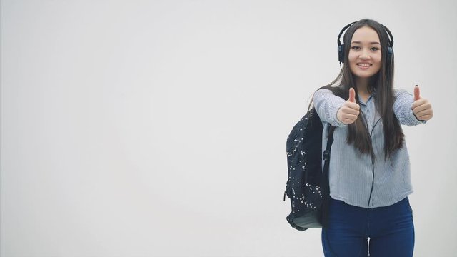 Inspired young asian girl in headphones appearing on the white background, backpack slung over her shoulder, giving thumbs up, smiling.