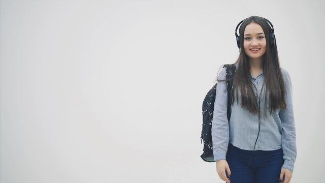 Inspired young asian girl in headphones, backpack slung over her shoulder, pointing fingers at the copyspace, giving thumbs up, smiling.