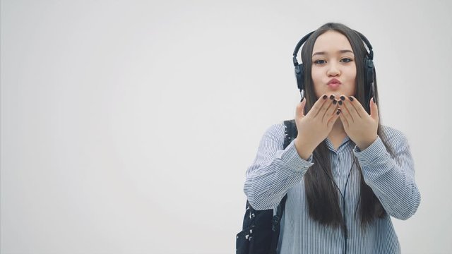 Pretty young asian girl in headphones backpack slung over her shoulder, giving an airkiss to the camera, smiling.