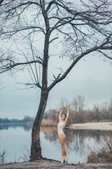 Ballerina dances at the lake shore near a tree.
