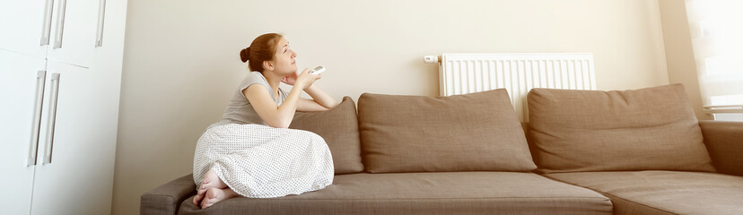 Young brunette woman in her modern apartment in living room. She turns on the air conditioner from the remote control sitting on the sofa. Climate control at home with split system, toned