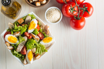 Fresh Caesar salad with delicious chicken breast, ruccola, spinach, cabbage, arugula, egg, parmesan and cherry tomato on wooden background. Oil, salt and pepper. Healthy and diet food concept.