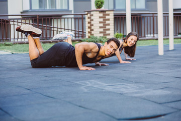 Naklejka premium Charming young blond girl and slender muscular man in red tops and black shorts doing an exercise plank on a lawn in a summer green park