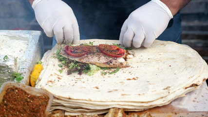 Traditional mediterranean cuisine. Man is making ekmek with fish, greens, tomatoes and seasonings at street market, hands in gloves closeup. Cook wraps the stuffing in pita.