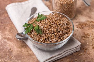 Cooked buckwheat in a glass, transparent bowl, parsley leaf, linen napkin, jars for storing cereals, brown background