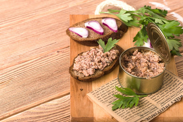 Canned fish, in an open tin, two pieces of dark bread on wooden cutting board, brown background