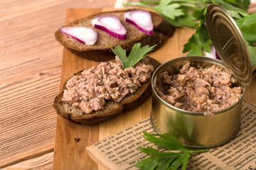 Canned fish, in an open tin, two pieces of dark bread, with onions and parsley, on a wooden cutting board