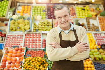 Waist up portrait of smiling senior man looking at camera while selling fruits and vegetables at...