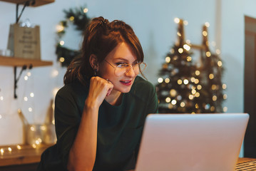 Smiling woman entrepreneur working on laptop sitting at home with christmas lights on the background