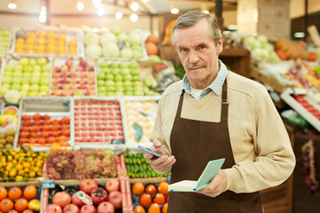 Waist up portrait of senior man holding notebook while selling fruits and vegetables at farmers market, copy space