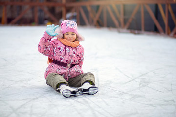 Girl shod in figure skates, sits on ice on a skating rink and waves a hand