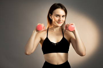 Athletic brunette girl in a black top with pink dumbbells and headphones on a gray background. Copy space