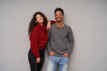 Indoor shot of pretty young brunette couple with dark skin posing over grey background in woolen cosy sweaters, looking cheerfully at camera and smiling broadly
