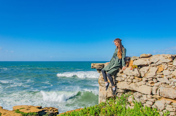 Obraz premium Woman dreamily looks at the Atlantic ocean and smiles, sits on the edge of a rocky shore, the wind blows hair, makes waves. Copy space.