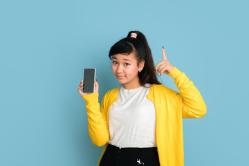 Asian teenager's portrait isolated on blue studio background. Beautiful female brunette model with long hair. Concept of human emotions, facial expression, sales, ad. Showing blank phone screen.