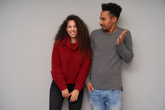 Cheerful attractive young curly lady with long dark hair looking happily at camera while young brunette bearded man with dark skin looking discontentedly at her and raising palm confusedly