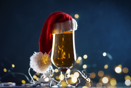 Festive Christmas beer glass in Santa hat on dark background with bokeh garland