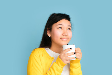 Asian teenager's portrait isolated on blue studio background. Beautiful female brunette model with long hair. Concept of human emotions, facial expression, sales, ad. Drinking coffee or tea.