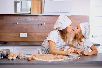 caucasian mother and child kissing while baking in kitchen, little girl helper. wearing aprons and cap
