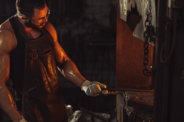 young strong muscular blacksmith wearing black apron and gloves for safety, stand preparing iron on fire, having powerful physique