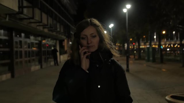 Girl with phone walk night city calling alone in dark park near the river in jacket and backpack cold autumn street lights low angle view