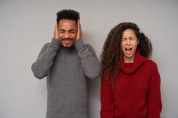 Portrait of young stressed curly brunette female screaming with wide mouth opened and keeping eyes closed while standing over grey background with dark skinned bearded man covering his ears with hands