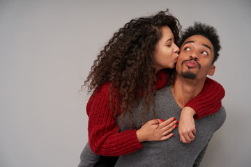 Obraz premium Portrait of young lovely long haired curly brunette woman embracing her smiling dark skinned bearded boyfriend and kissing him gently on cheek, isolated over grey background