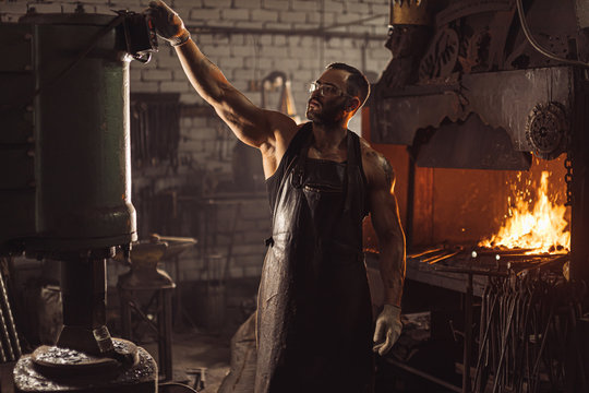 muscular skillful forger at workshop wearing leather apron isolated in dark space, furnace in workshop