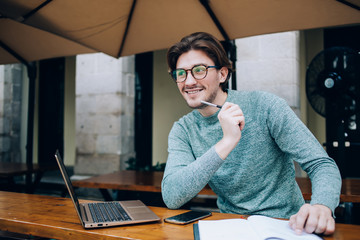 Cheerful man contemplating while sitting with notepad and laptop in street cafe