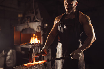 young caucasian forger beating hot metal using hammer isolated in dark space workshop, performing blacksmith work