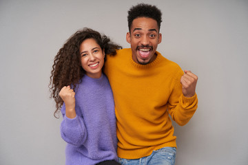 Fototapeta premium Portrait of cheerful young pretty dark skinned brunette pair looking happily at camera and raising fists joyfully, rejoicing together about nice occasion, isolated over grey background
