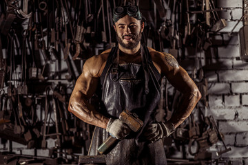 Obraz na płótnie Canvas portrait of young smiling muscular strong caucasian man blacksmith looking at camera, wearing leather apron uniform, holding hammer isolated in dark space, workshop