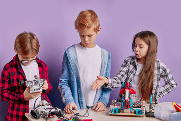 Fototapeta premium portrait of enthusiastic three kids working as team, assembling robots. three future engineers look at table full of robotics, two boys and one girl