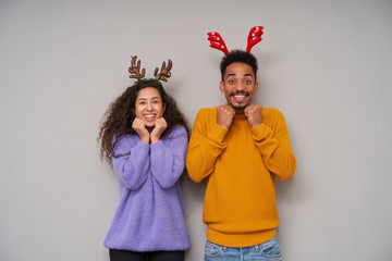 Overjoyed lovely young dark skinned curly couple looking happily at camera with wide smile and keeping raised fists under their chins, wearing holiday hoops and woolen sweaters over grey background