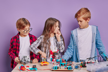 Portrait of caucasian little children assembling robots - group of happy kids or students with electric toys and building robots at robotics school lesson. isolated over purple background