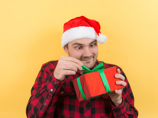 Pensive man in a red hat, holds a gift with interest on his face. Positive human emotions for christmas, secret santa. Yellow background, copy space.