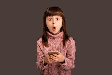 Shocked by an unexpected surprise, a girl on a gray background is holding a mobile phone in her...