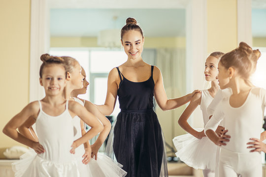 Young Trainer Of The Ballet School Helps Young Ballerinas Perform Different Choreographic Exercises In Studio. Children Girls Rehearse Together In The Ballet Class