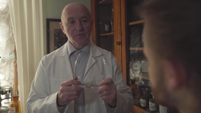 Portrait of baldheaded senior man in white robe taking off eyeglasses and talking to blurred man at the foreground. Old Caucasian pharmacist educating new worker in old-fashioned drugstore.