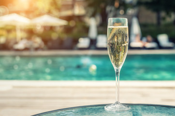 Close-up white champagne or prosecco glass against poolside at luxury resort hotel during vacation. Sparkling wine with rising bubbles with blue pool background outdoor. Refreshing alcohol drink
