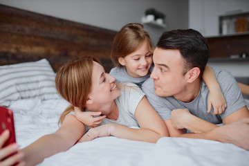 emotional happy parents lying on bed together with daughter using red smartphone, indoors. portrait