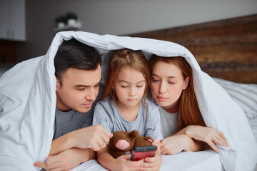 happy caucasian family use smartphone while spending time together at home under blanket , watching video. leisure time