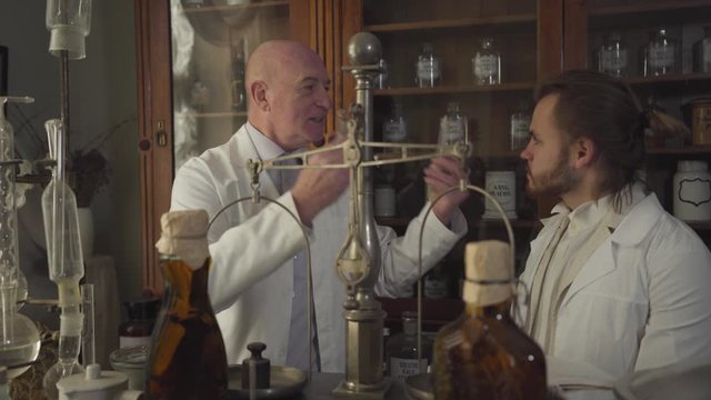 Happy old Caucasian man showing vintage test tube and flask to young man and talking. Apothecaries in white robes working in ancient pharmacy. 19th century, vintage, retro drugstore.