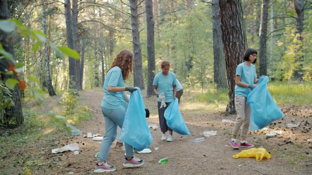 Students girls and guys are cleaning forest on Earth day collecting trash in plastic bags busy with charity work on sunny day. Youth and volunteering concept.