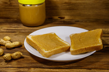 Jar of peanut butter and plate with two sandwiches with peanut butter on wooden table